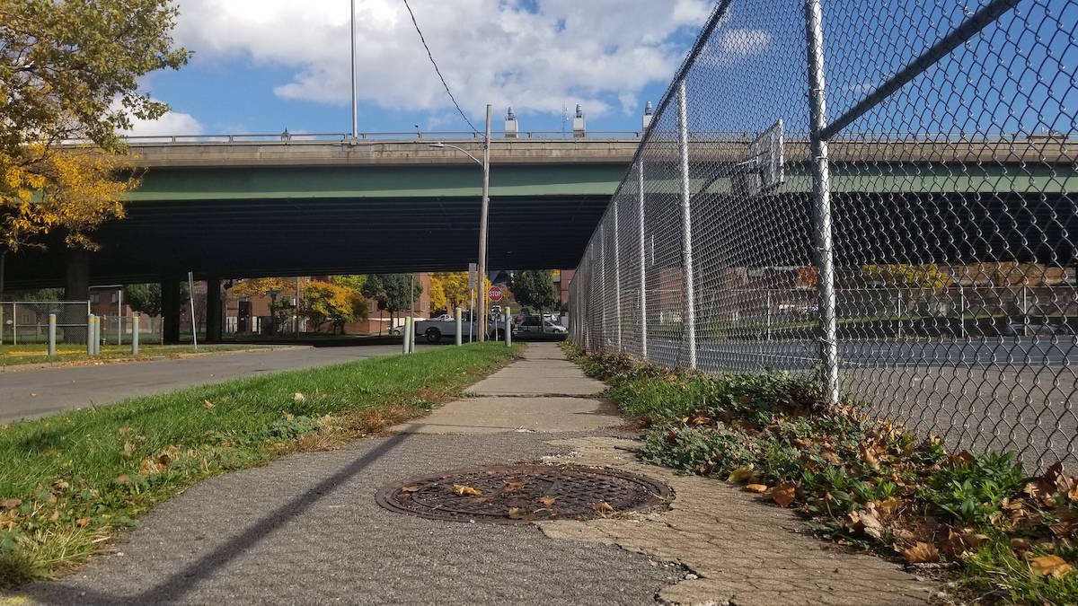 A cracked sidewalk and chainlink fence leads under the I-81 viaduct, which creates shadows at the end of the path.