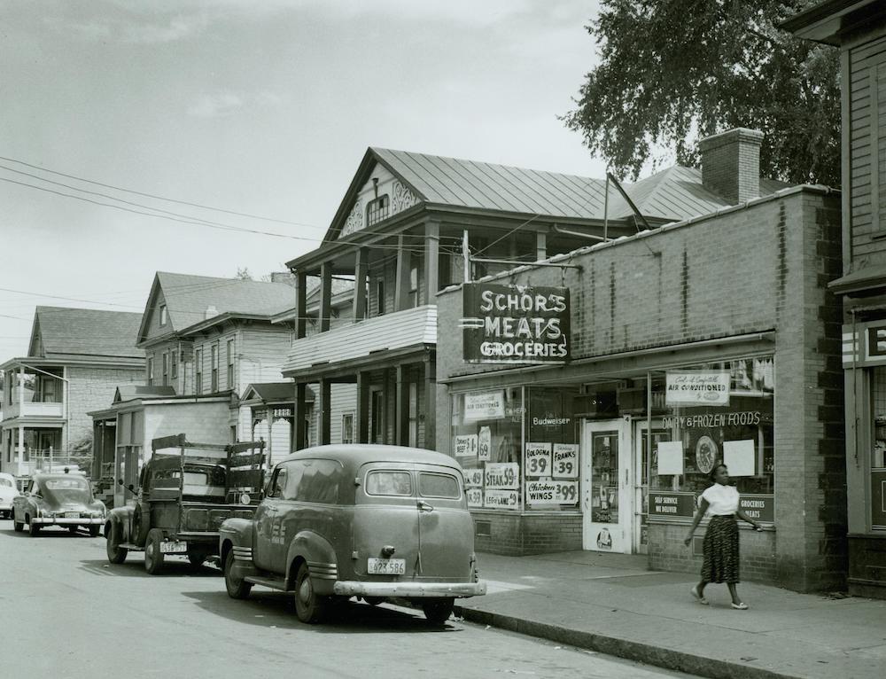 A woman in an ankle-length skirt walks past a single-story building with the sign “Schor’s Meats Groceries” next door to several wood-frame residential houses. 
