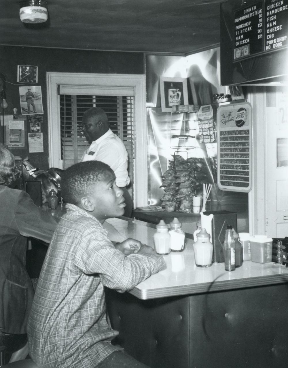 A young boy contemplates what to order at a lunch counter as an employee works in the background. 