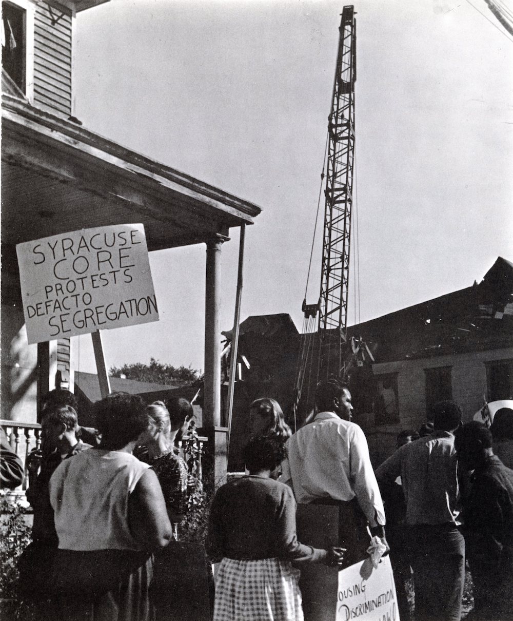 A group of protesters gather in front of a construction crane, with one protester holding up a sign that says “Syracuse CORE protests defacto segregation” and another holding a sign that says “housing discrimination” and is cut off at the bottom of the photo.  