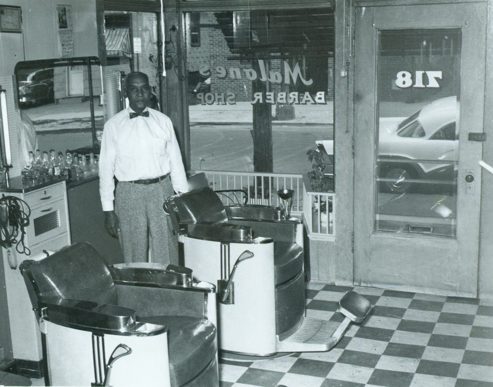 A man wearing a white button-up shirt and a black bowtie stands behind a barbershop chair. A sign in the window behind him reads “Malone’s Barber Shop.”