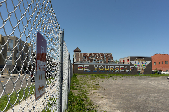 A fenced off gravel lot with the tarp-covered roof of the former church building at 711 East Fayette Street in the distance, and a mural on the side of the XO Taco building that reads “Be Yourself.” 