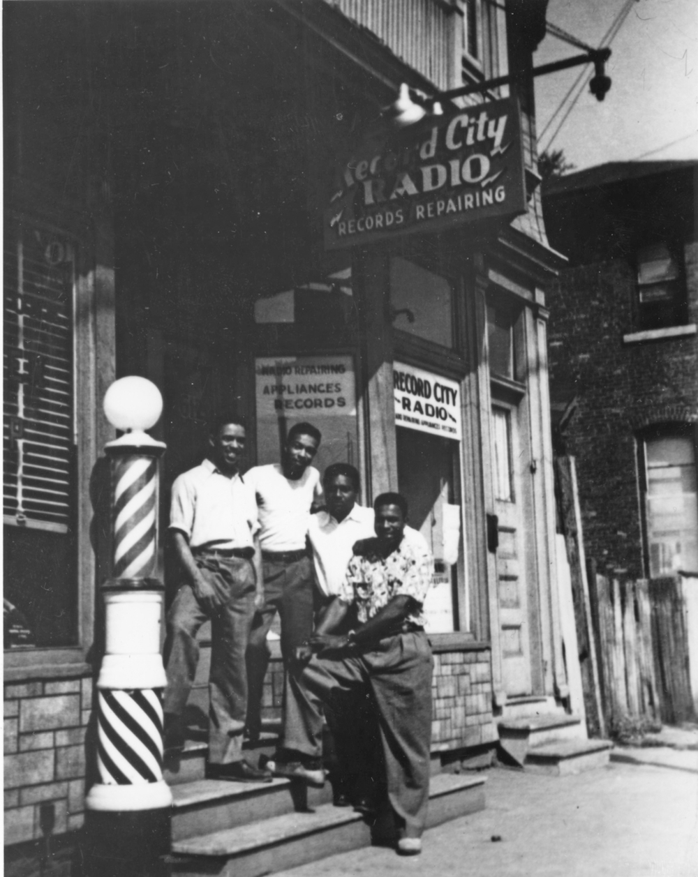Four men stand together on the steps in front of a barbershop. The sign to the shop next door reads “Record City Radio Records Repairing.”