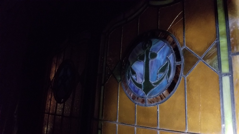 On the backdrop of orange stained-glass window panels, a blue circle of stained glass in the center features the symbol of an anchor.