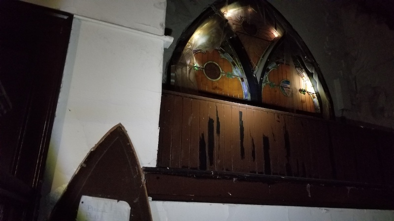 The church’s main gothic-arched window contains three segments of stained glass. Though it is dark in the church and the windows are boarded, a few spots of light stream in from around the window’s edges. 