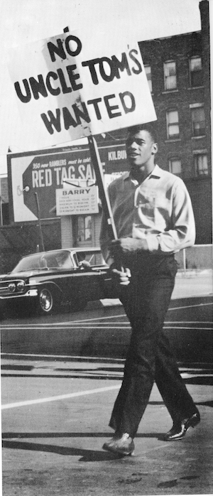 A young man wearing suit pants and a button up shirt carries a sign that says “No Uncle Toms Wanted.”